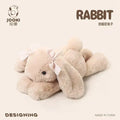 Light beige plush rabbit pillow, ribbon bow on body, lying with legs forward.