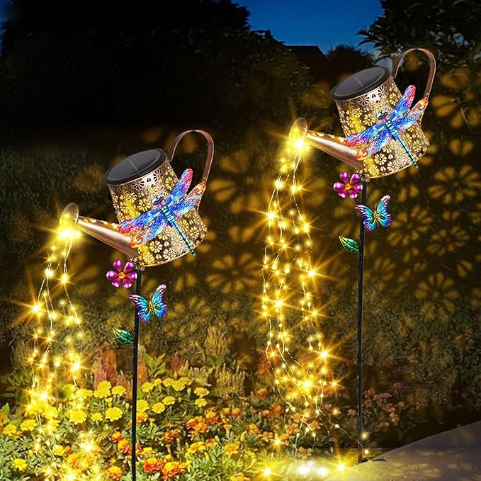 Two solar watering can stake lights with dragonfly decorations, warm yellow glow outdoors