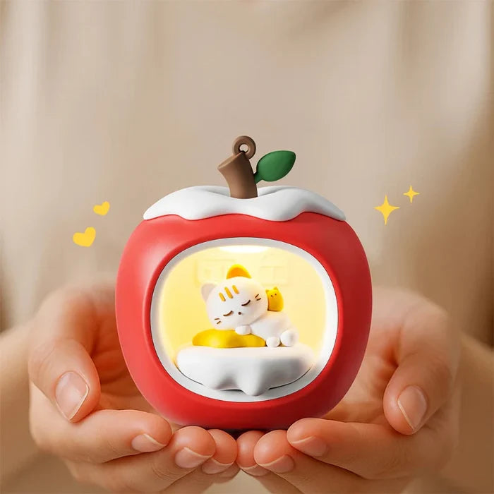 Apple-shaped night light held in hands, red body and warm-lit window with sleeping cat.