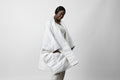 A woman wearing a white Blanket Robe with hands in side pockets, standing against a light background