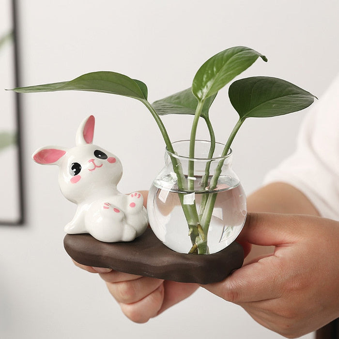 Ceramic bunny planter beside a clear glass vase with green leaves, mounted on dark base