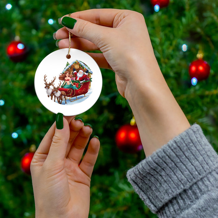Round ceramic ornament with mermaid and Santa sleigh scene held by hand on a Christmas tree