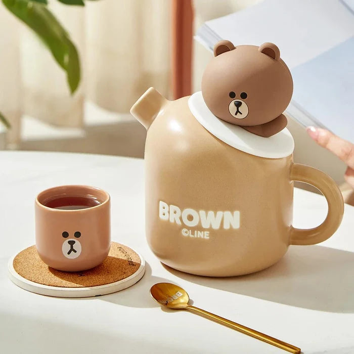 Ceramic teddy bear teapot with handle, brown cup, and spoon; bear-face logo on front.