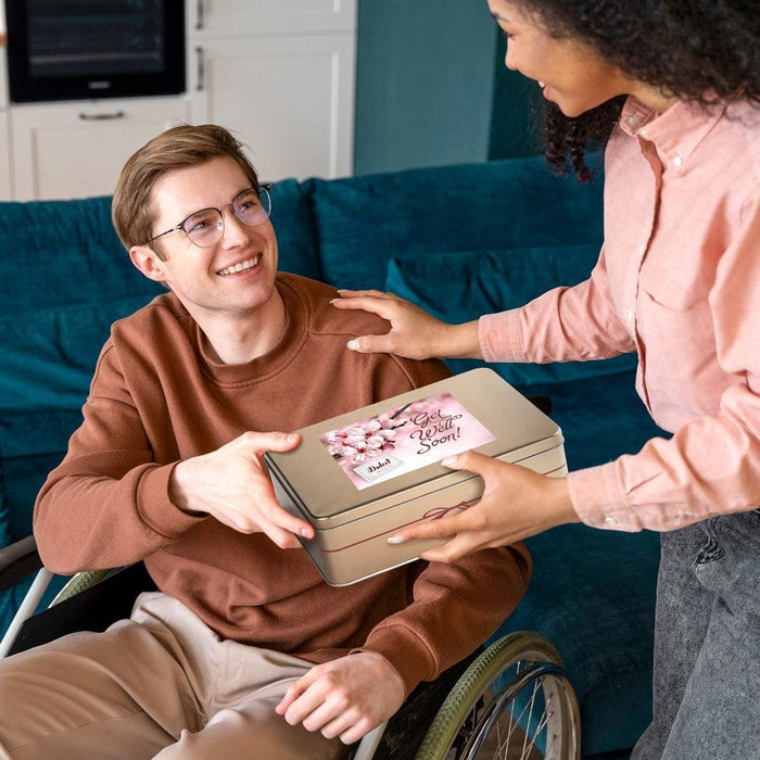 Person holding a cookie gift tin with “Get Well Soon” label in a living room