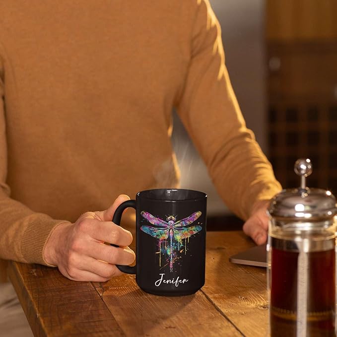 Black ceramic coffee mug with colorful dragonfly graphic and custom name on wooden table