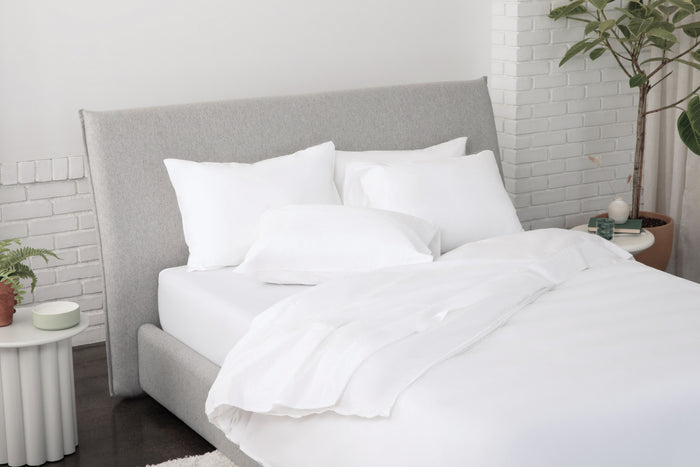 White bedding set on a bed with a gray headboard in a room with plants and a white wall.