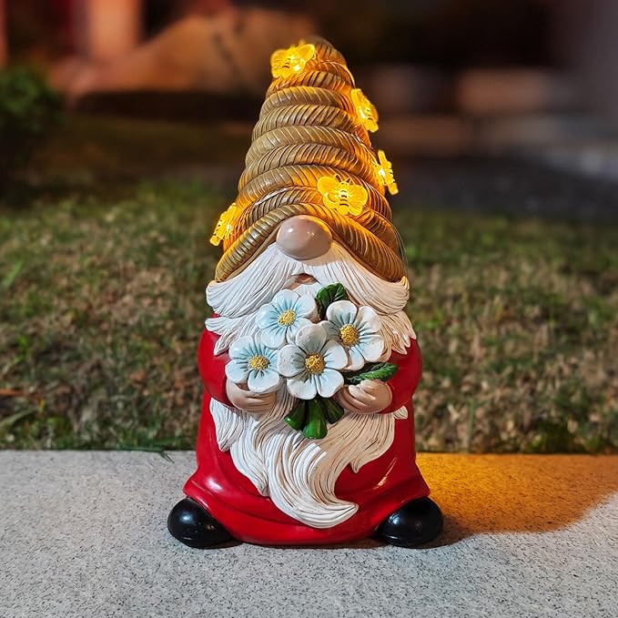 Bee-themed solar LED garden gnome statue standing on pavement, red coat, white beard, blue flowers, lit