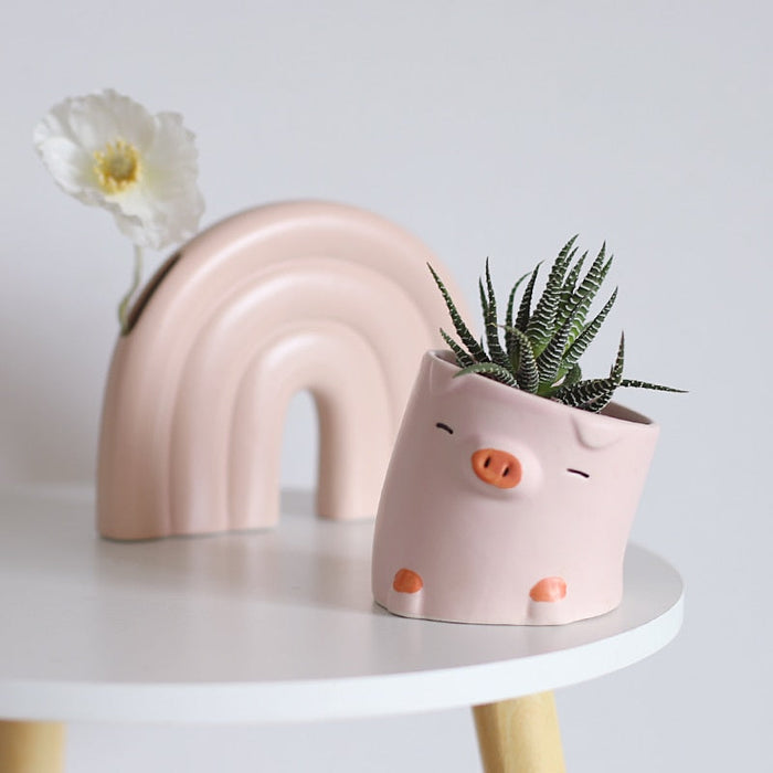 Pink pig ceramic planter with succulent in front of a pastel rainbow arch planter.