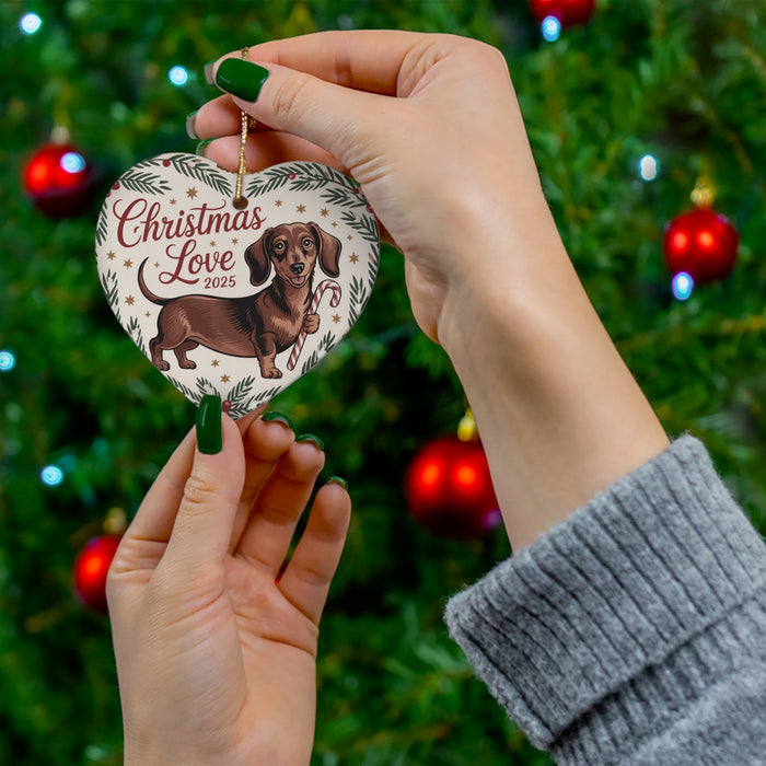 Hands hold heart-shaped dachshund ceramic ornament reading “Christmas Love 2025”.