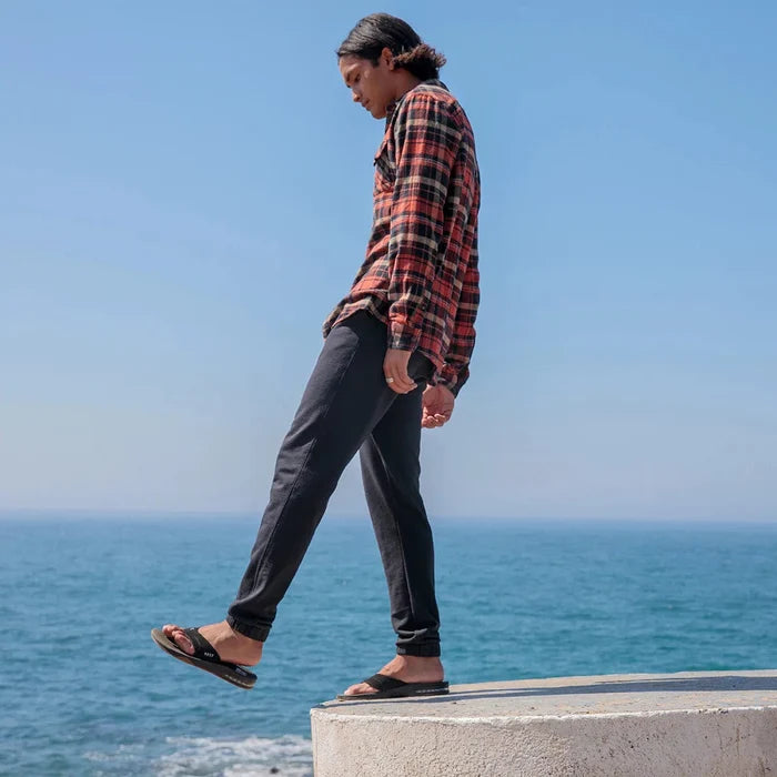 Reef Fanning Flip Flops in black with cushioned footbed and bottle opener on sole, worn outdoors by the ocean.