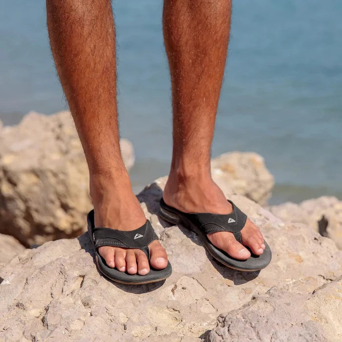Reef Fanning Flip Flops in black with padded straps and signature arch support, worn outdoors on rocky terrain by the ocean.