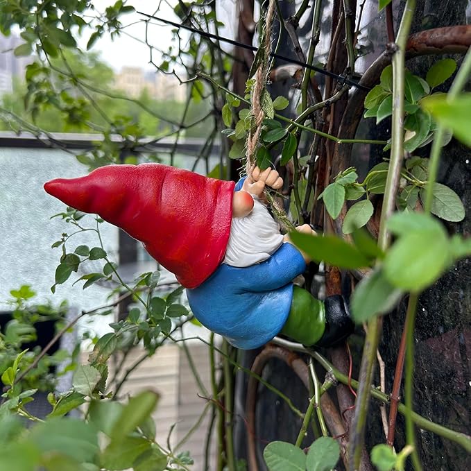 Climbing gnome garden decoration on rope among green leaves, red hat, white beard, blue body