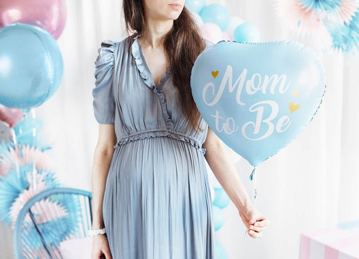 Blue heart balloon with “Mom to Be” text, held near a baby shower outfit.