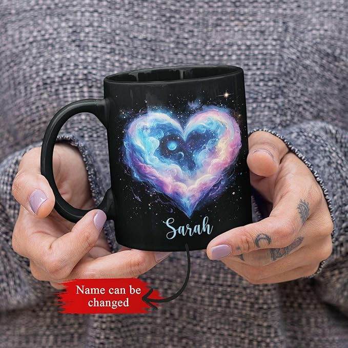 Black ceramic mug held in hands, featuring galaxy heart design and name Sarah