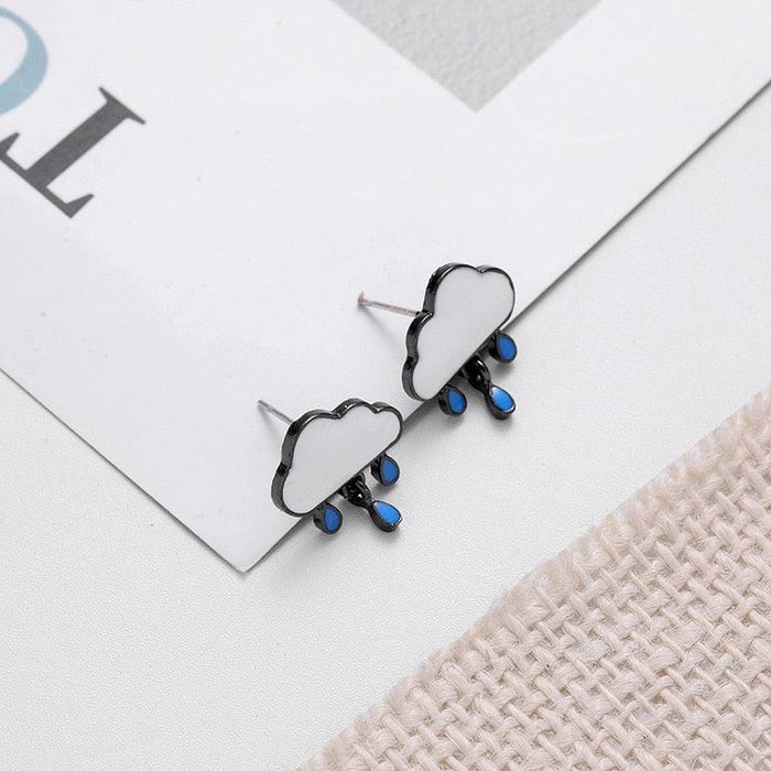 Cloud stud earrings with blue raindrops and black outline, lying on paper