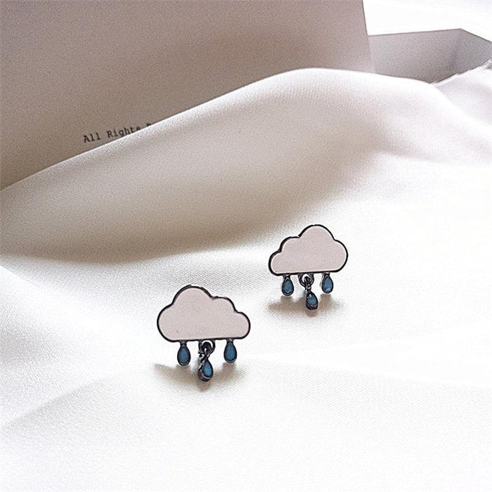 Cloud rain drop earrings with blue drops, white enamel, shown on soft bedding