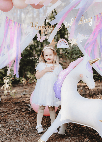 Unicorn balloon next to a child at a pastel birthday balloon arch